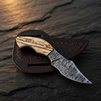 A professional handmade Damascus EDC cowboy knife resting on its leather pancake sheath, showcasing the ashwood handle, copper pins, and patterned blade on a dark stone surface