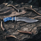 A professional handmade Damascus Bowie knife featuring a 10-inch San Mai VG10/15N20 steel blade and a unique blue resin handle, artistically displayed on a dark stone background with antler accents.