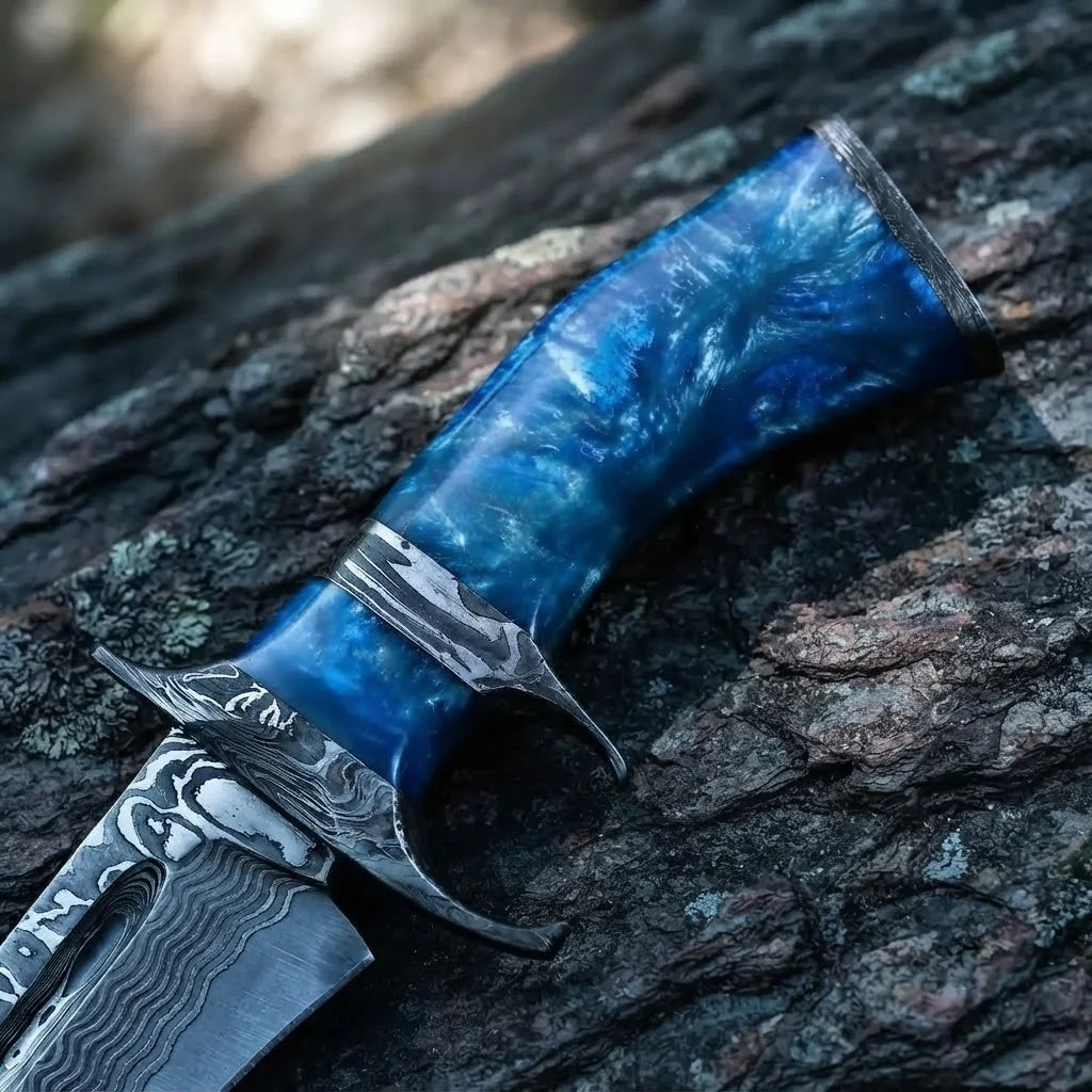 Close-up of a handmade Damascus Bowie knife, showcasing the striking blue swirl resin handle, Damascus steel finger guard, and bolster against a dark rugged stone background