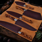 A professional set of hand-forged Damascus steel kitchen knives with intricate floral wooden handles, each paired with a custom-fitted maroon leather sheath on a rustic surface.