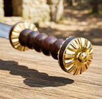 A professional macro close-up of a handmade Damascus Roman Gladius sword showcasing the intricately fluted brass pommel and ridged wooden handle resting over a wooden surface outdoors