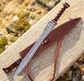 A professional top-down outdoor studio shot of a handmade 24-inch Damascus steel Roman Gladius sword laid diagonally across its custom leather scabbard and straps on a rustic wooden bench under natural sunlight