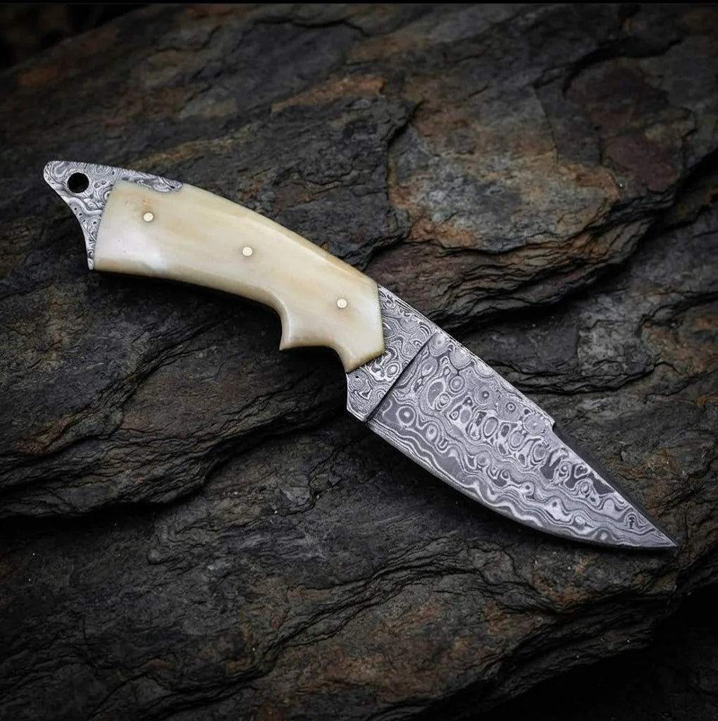 A close-up view of a handmade Damascus steel skinner knife showcasing its intricate 512-layer blade pattern and smooth camel bone handle, resting on a dark stone surface