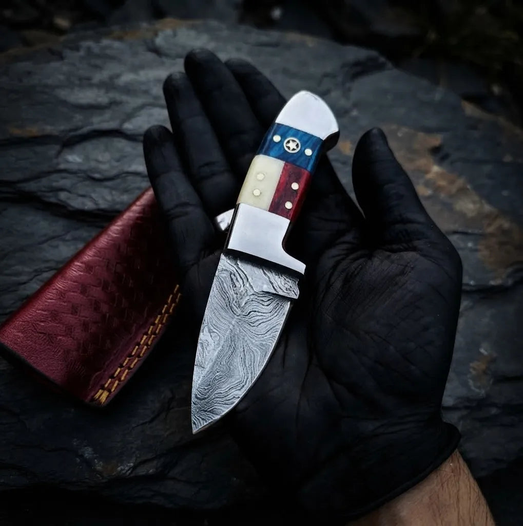 A close-up hand-held view of a handmade Damascus steel skinner knife featuring a unique American flag handle, perfect for EDC and hunting tasks.