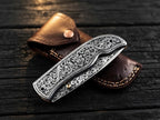 High-quality custom engraved pocket knife showcasing complex metal carving in a compact folded position alongside an artisan leather carry case