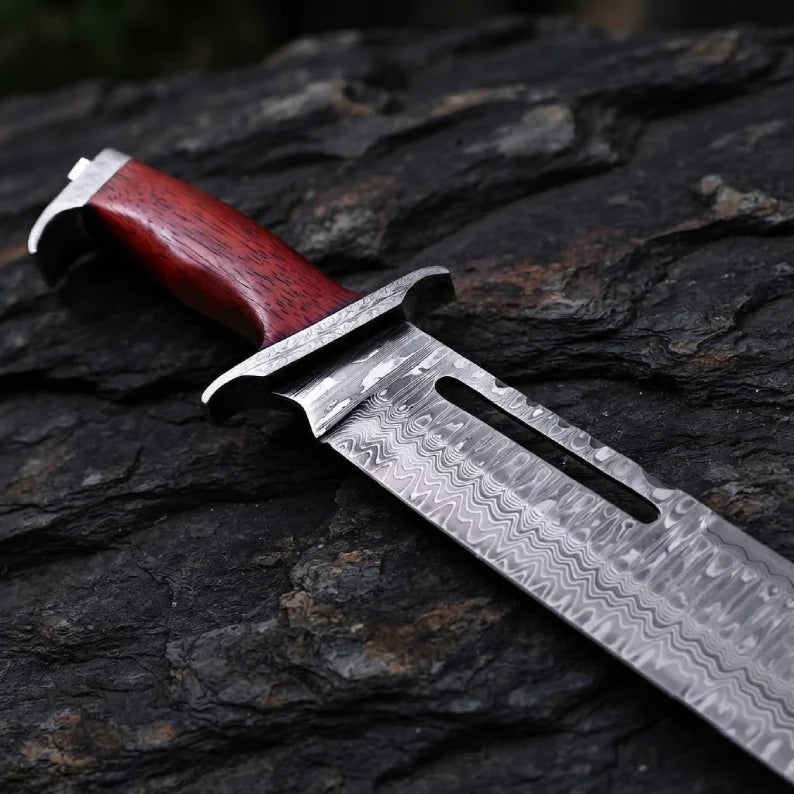 Close-up of a handmade Rambo III style Bowie knife, showcasing the ergonomic Pakka wood handle, solid bolster, and intricate wavy Damascus steel pattern on the blade.