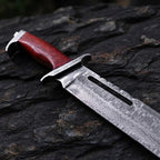 Close-up of a handmade Rambo III style Bowie knife, showcasing the ergonomic Pakka wood handle, solid bolster, and intricate wavy Damascus steel pattern on the blade.