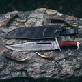 Handmade Rambo III style Bowie knife featuring a premium VG10 Damascus steel blade and ergonomic Pakka wood handle, displayed with a leather sheath on a stone background.