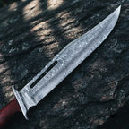 Close-up of a handmade Rambo III style Bowie knife, showcasing the intricate wavy Damascus steel pattern and distinct central fuller on a rugged stone background