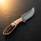 A professional studio shot of a handmade Western belt knife featuring a curved D2 rasp steel blade, copper bolster, and genuine stag horn handle on a dark stone background