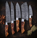 A set of luxury hand-forged Damascus steel kitchen knives featuring unique wavy blade patterns and intricate, hand-carved wooden handles, arranged with cinnamon and bay leaves