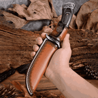 A person holding a handcrafted Damascus knife inside a premium brown leather sheath with embossed scallop borders, displayed against a rustic wood and dry leaf background.