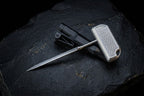 A minimalist titanium push dagger featuring a secure pocket clip for EDC carry, resting on a textured dark surface.