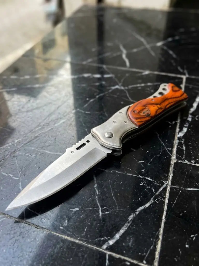 An angled perspective studio shot of an open handcrafted folding pocket knife on a black marble counter, focusing on the sharp satin-finish stainless steel blade with spine jimping, a solid steel bolster, and a contoured handle featuring a polished orange-brown wood inlay.