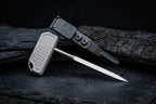 Textured titanium T-handle push dagger inserted into a black custom sheath with an attached UltiClip, resting on a dark volcanic rock with a tree trunk background for rugged EDC showcase.