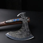 Extreme close-up of a handcrafted Viking bearded axe head resting on a dark surface, highlighting intricate runic engravings across the weathered metal blade and silver metal accents with inlaid blue stones near the wooden handle.