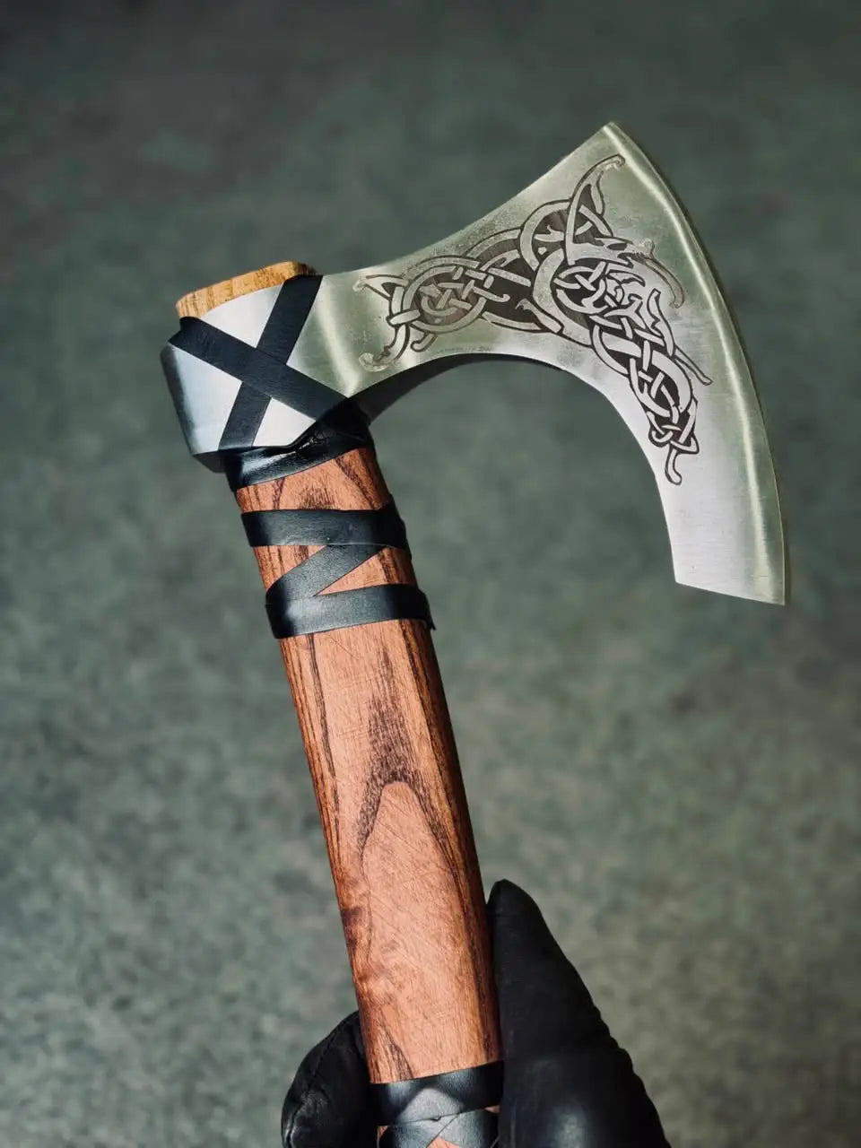 Close-up shot of a hand wearing a black leather glove holding a handcrafted Viking bearded axe, showcasing the intricate Norse knotwork engraving on the polished steel blade and a rich wood grain handle with black leather wrapping.