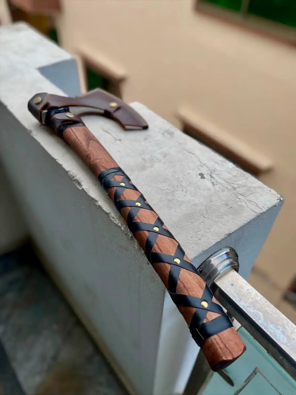 High-angle outdoor shot of a handcrafted Viking axe resting on a concrete ledge, showcasing a smooth wooden handle with a black leather criss-cross wrap secured by brass pins, and a brown leather protective sheath covering the blade.