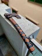 High-angle outdoor shot of a handcrafted Viking axe resting on a concrete ledge, showcasing a smooth wooden handle with a black leather criss-cross wrap secured by brass pins, and a brown leather protective sheath covering the blade.