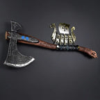 Flat lay of a handcrafted Viking bearded axe with runic blade engravings, blue accents, and a carved wooden handle, displayed horizontally next to a small brass Viking longship model on a dark grey surface.
